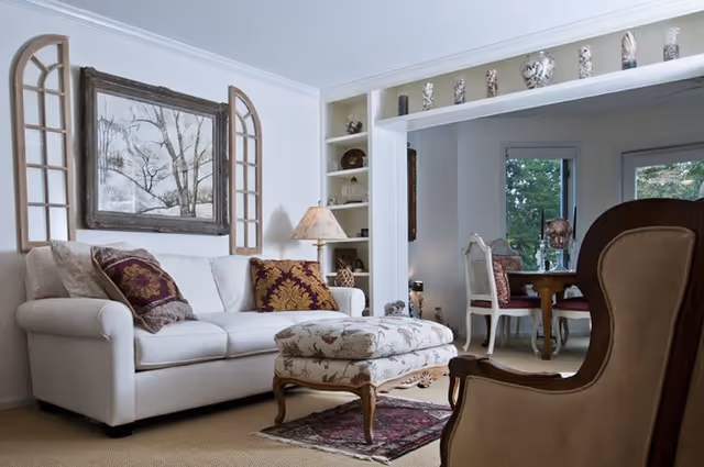 A cozy living room with a white sofa adorned with decorative pillows, a floral upholstered ottoman on a small rug, a wooden armchair in the foreground, and a lamp on a side table. The room opens into a dining area with a wooden table and chairs near windows showing greenery outside. Shelves with decorative items are built into the wall.