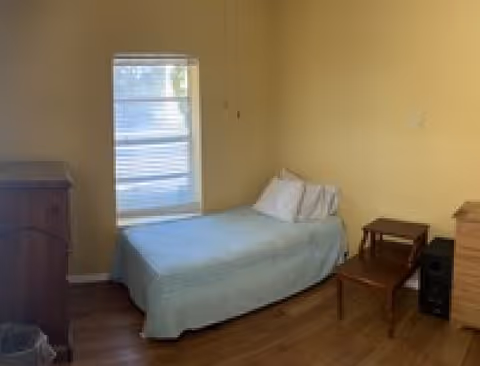 A small bedroom with a single bed covered with a light blue blanket and two white pillows. There is a window with blinds on the wall behind the bed. The room has wooden flooring and light yellow walls. Next to the bed are two wooden side tables stacked on top of each other and a wooden dresser on the right side. Another wooden piece of furniture is partially visible on the left side.