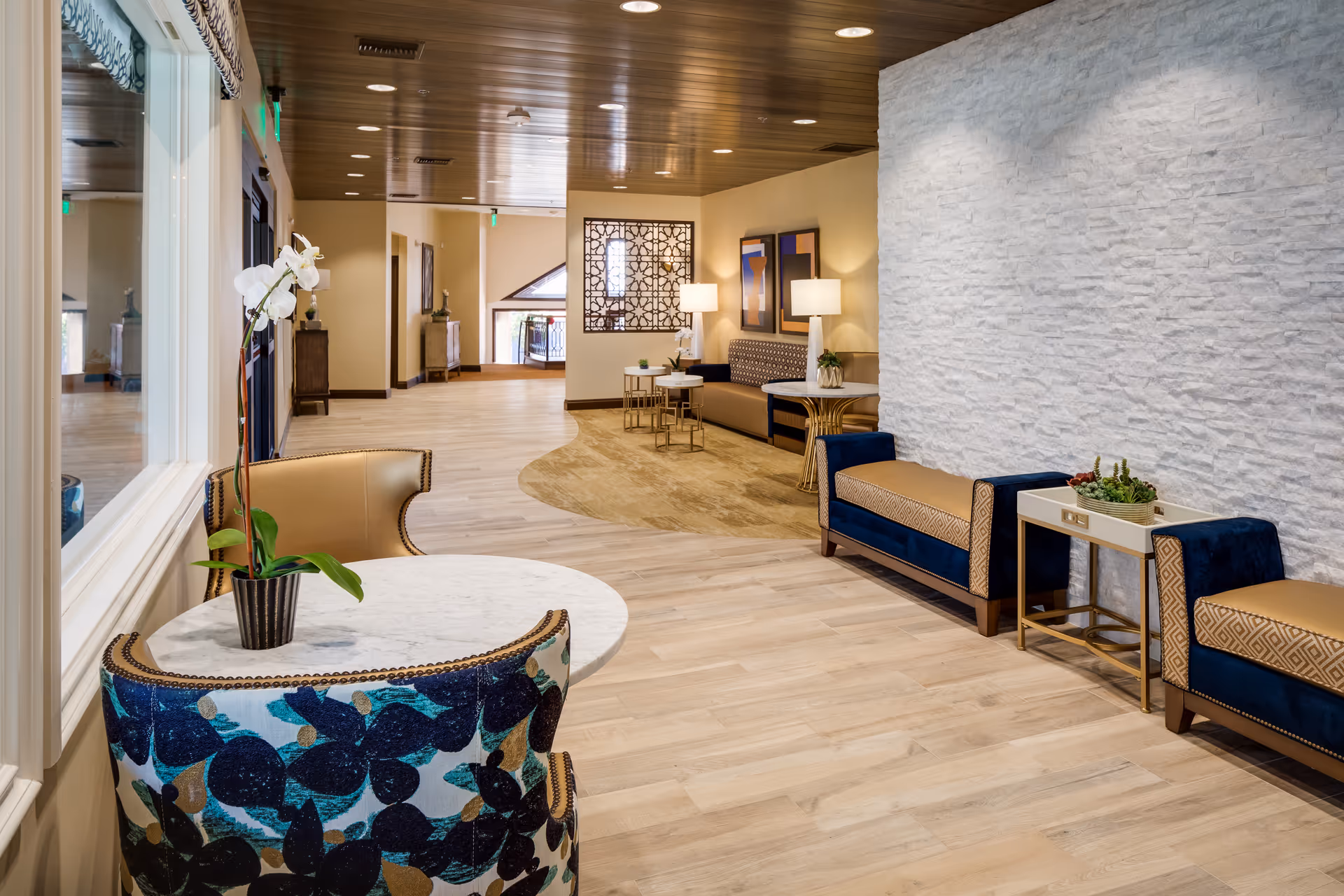 A bright, modern seating lounge with upholstered chairs and benches, small tables, potted plants, and decorative wall panels.