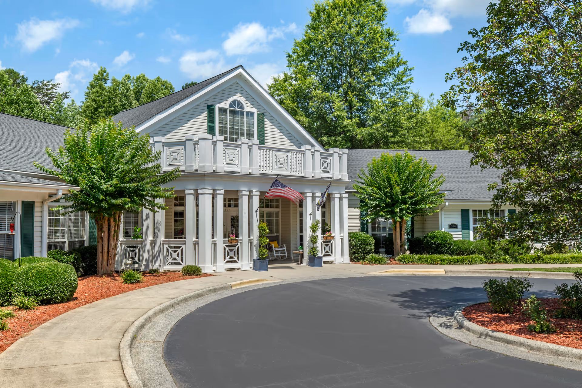 Front exterior view of Brookdale Asheboro facility with a curved driveway, white columns, American flag, and well-maintained landscaping including trees and bushes under a blue sky with scattered clouds.