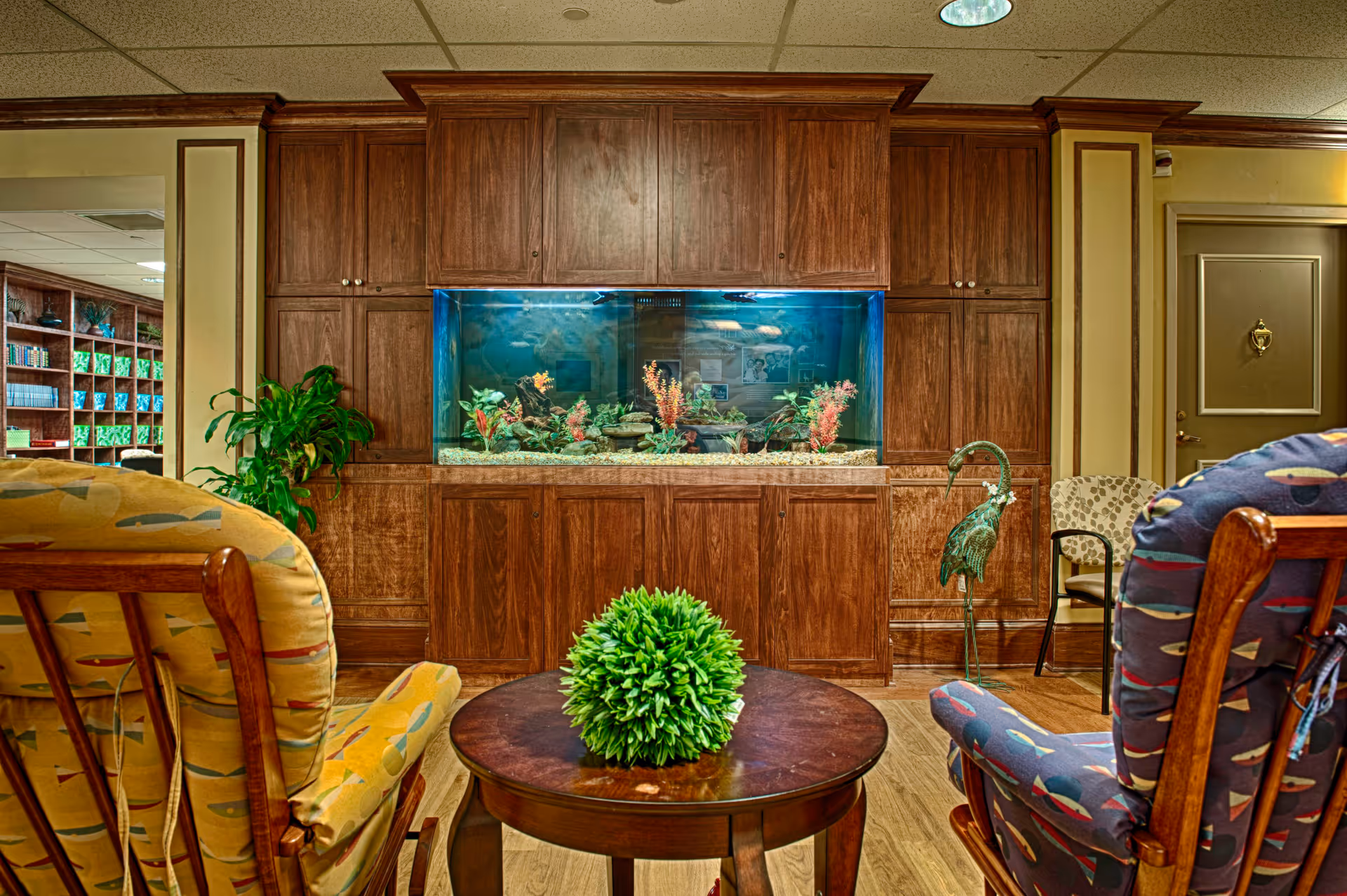 Seated lounge area with upholstered rocking chairs facing a built-in aquarium set in wooden cabinetry and a round coffee table topped with a green plant.