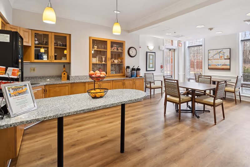 A bright dining area in an assisted living facility featuring a granite countertop island with a two-tiered fruit basket holding apples and oranges. Behind the island, there are wooden cabinets with glass doors displaying dishes and decorative items. To the right, there are several tables with chairs arranged near large windows with blinds, allowing natural light to fill the room. The walls are decorated with framed artwork and a clock.