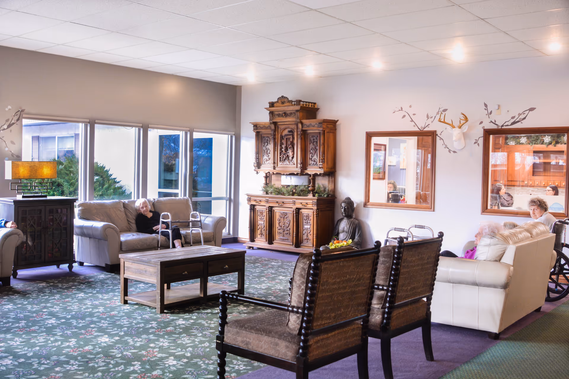 A bright and spacious living room in an assisted living facility with several elderly residents seated on sofas and chairs. The room features large windows letting in natural light, a wooden coffee table, an ornate wooden cabinet, a Buddha statue, and decorative wall art including a mounted deer head. The carpet has a floral pattern, and the overall atmosphere is calm and comfortable.