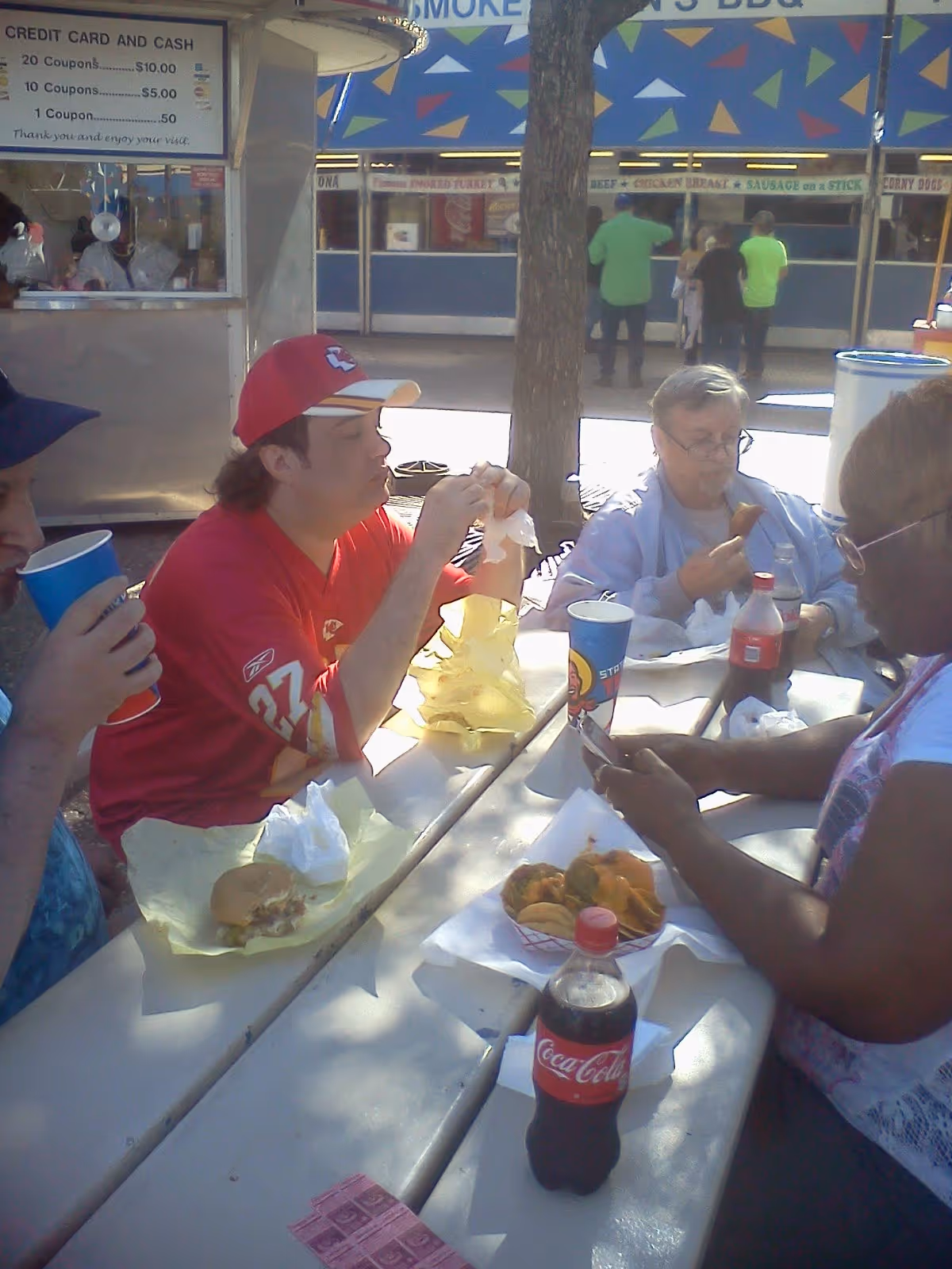 Four people sitting at a picnic table outdoors eating food and drinking soda. There are food stands in the background with signs for various foods and prices. One person is wearing a red sports jersey and cap, another is using a smartphone, and there are cups and food wrappers on the table.