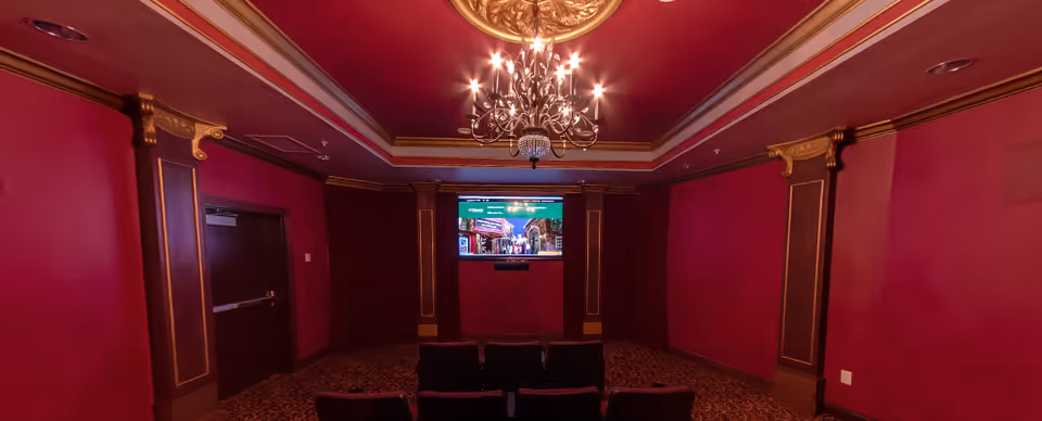 A small theater room with red walls and ornate gold trim. There is a chandelier hanging from the ceiling, a large screen mounted on the front wall, and several rows of black chairs facing the screen.