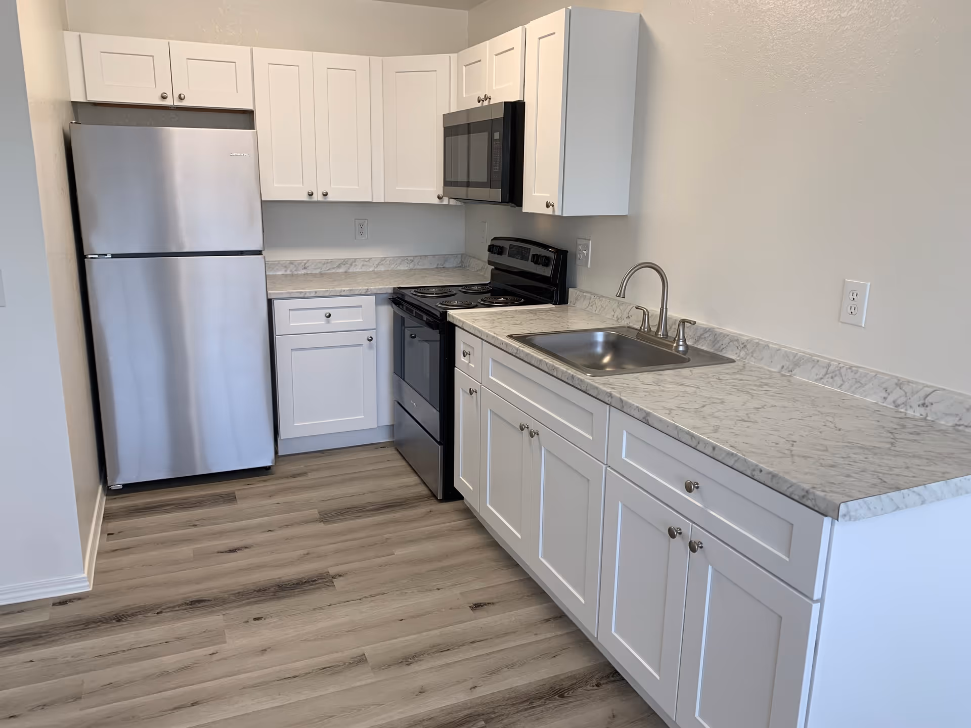 A modern kitchen with white cabinets, a marble-patterned countertop, a stainless steel refrigerator, an electric stove with oven, a microwave above the stove, and a stainless steel sink with a faucet. The floor is light wood laminate.