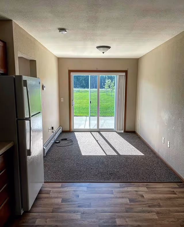Interior view of a small dining area with carpeted floor next to a kitchen with wood flooring and a refrigerator on the left. The dining area has a sliding glass door leading to a green outdoor space with grass and trees. The walls are beige, and there is a ceiling light fixture above the dining area.