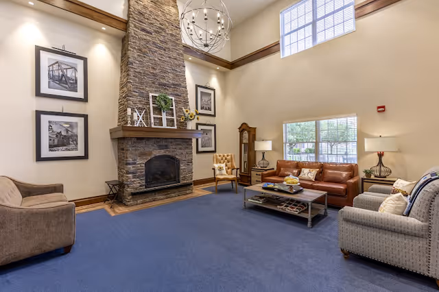 A spacious living room with a high ceiling and large windows letting in natural light. The room features a stone fireplace with decorative items on the mantel, including framed pictures and a small plant. There are several seating options including a brown leather sofa, a beige armchair, a patterned armchair, and a tufted chair. A coffee table with magazines and decorative items sits in the center on a blue carpet. The walls are adorned with black and white framed photographs, and two table lamps provide additional lighting.