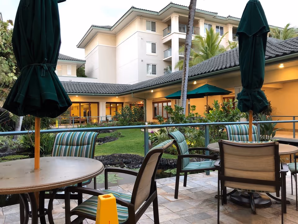 Courtyard patio with round tables, striped chairs and closed green umbrellas overlooking landscaped grounds and a multi-story building.