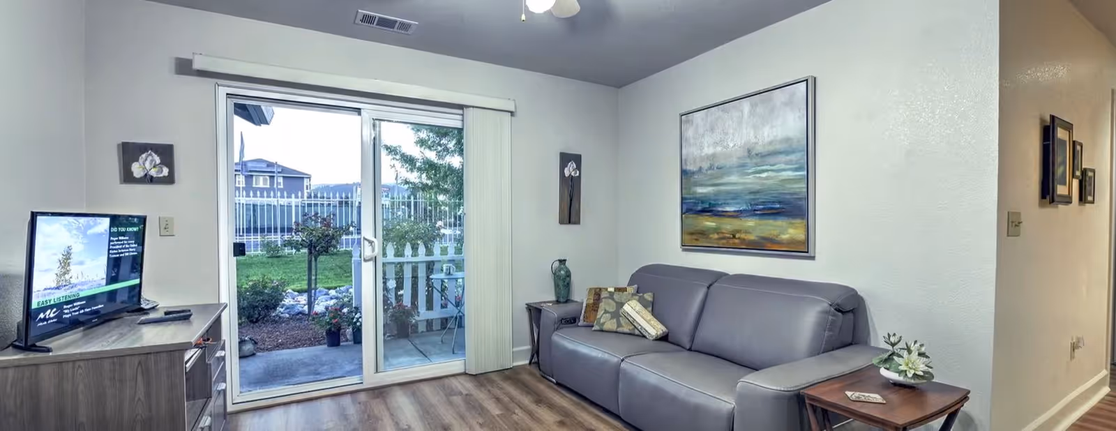 A cozy living room with a gray leather sofa adorned with decorative pillows, a wooden side table with a small flower arrangement, and a large abstract painting on the wall. Opposite the sofa is a wooden TV stand with a flat-screen television. Sliding glass doors lead to an outdoor patio with a white picket fence and greenery visible outside. The room has light-colored walls and wood flooring.