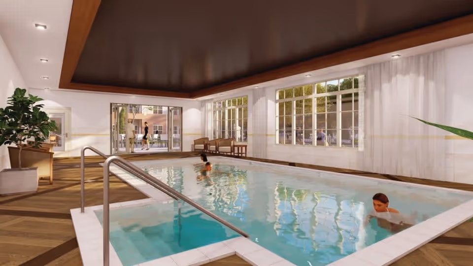 Indoor swimming pool area with wooden flooring and large windows letting in natural light. Two people are swimming in the pool. There are chairs and plants around the pool, and a door leading to an outdoor area where two people are standing.