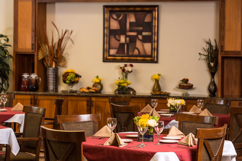 A dining room with tables covered in red and white tablecloths, set with plates, napkins, wine glasses, and floral centerpieces. In the background, there is a wooden cabinet with decorative vases, fruit bowls, and a framed abstract painting on the wall.