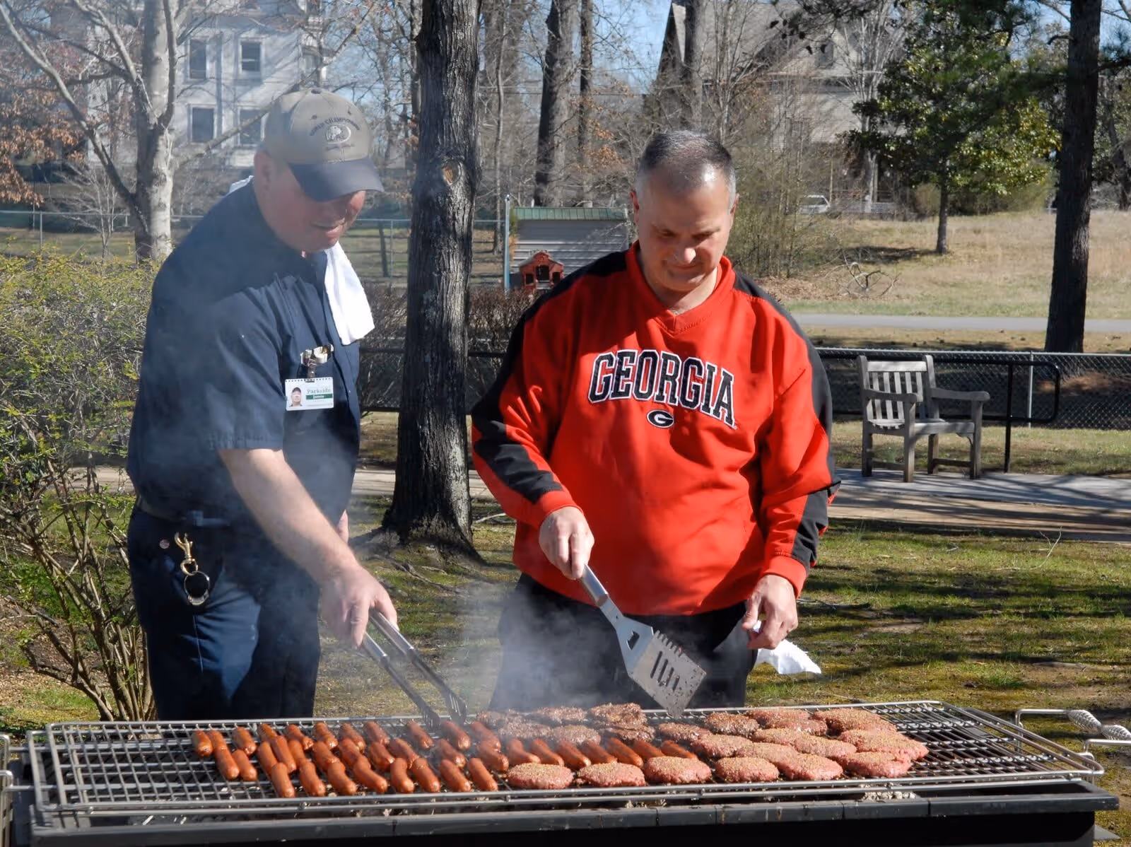 Two men grilling hot dogs and hamburgers outdoors on a large barbecue grill. One man is wearing a red Georgia sweatshirt and using a spatula, while the other man is wearing a dark shirt and cap with a towel over his shoulder, holding tongs. Trees, a bench, and a house are visible in the background.