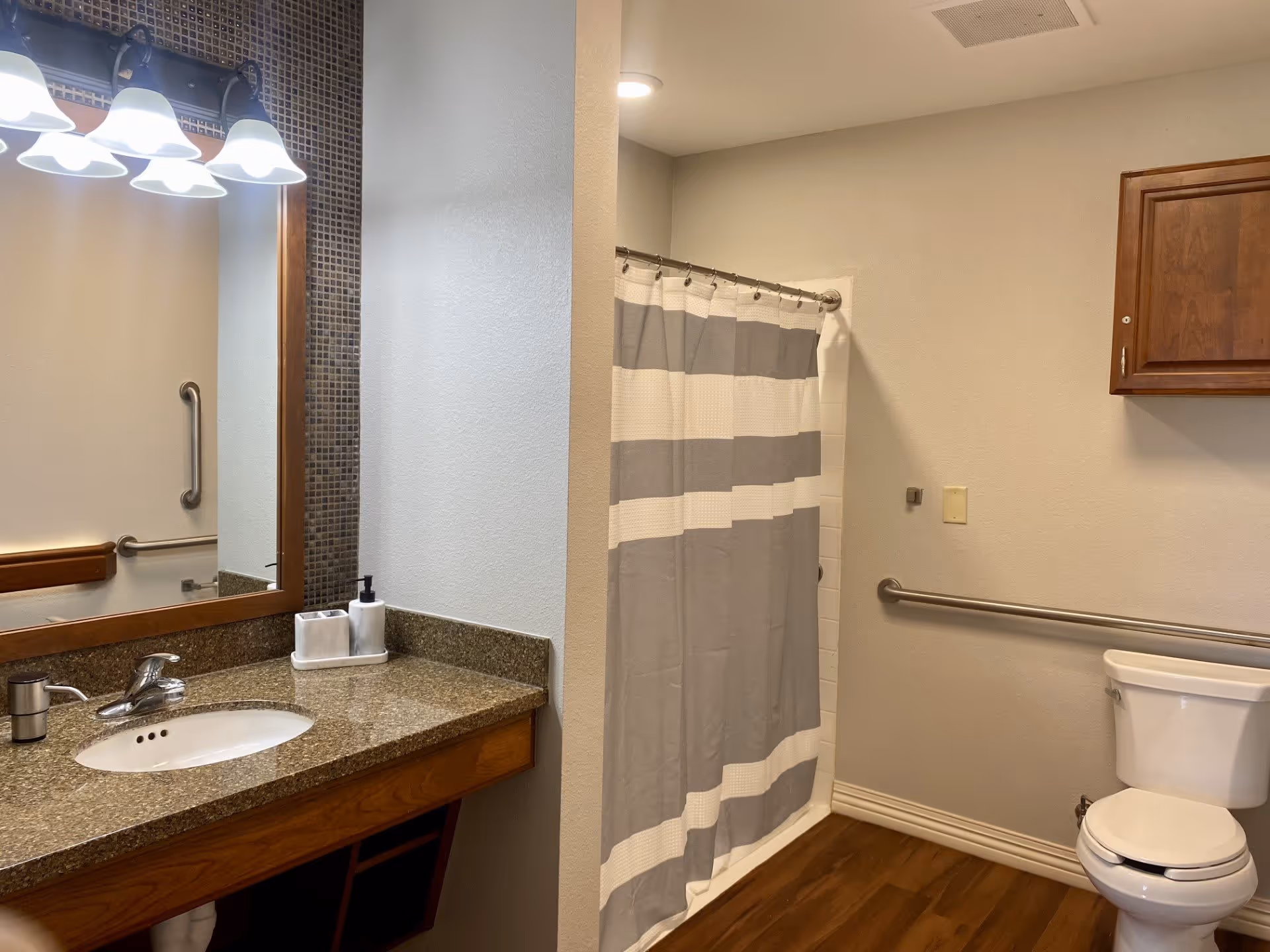 Bathroom with a granite countertop sink, a large mirror with mosaic tile border, a light fixture with four lights above the mirror, a shower with a gray and white striped curtain, a toilet, wooden cabinet mounted on the wall, and grab bars for accessibility.