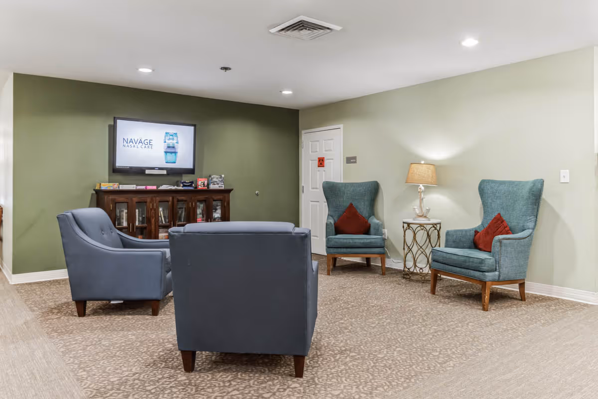 A senior living facility lounge with several armchairs arranged around a TV mounted on a green accent wall, and a side table with a lamp.