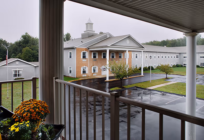 View from a covered balcony overlooking a wet parking lot and a large two-story building with white columns and a cupola on the roof. There are some small trees and green grass around the building, and a flower pot with orange and yellow flowers is visible on the balcony railing.