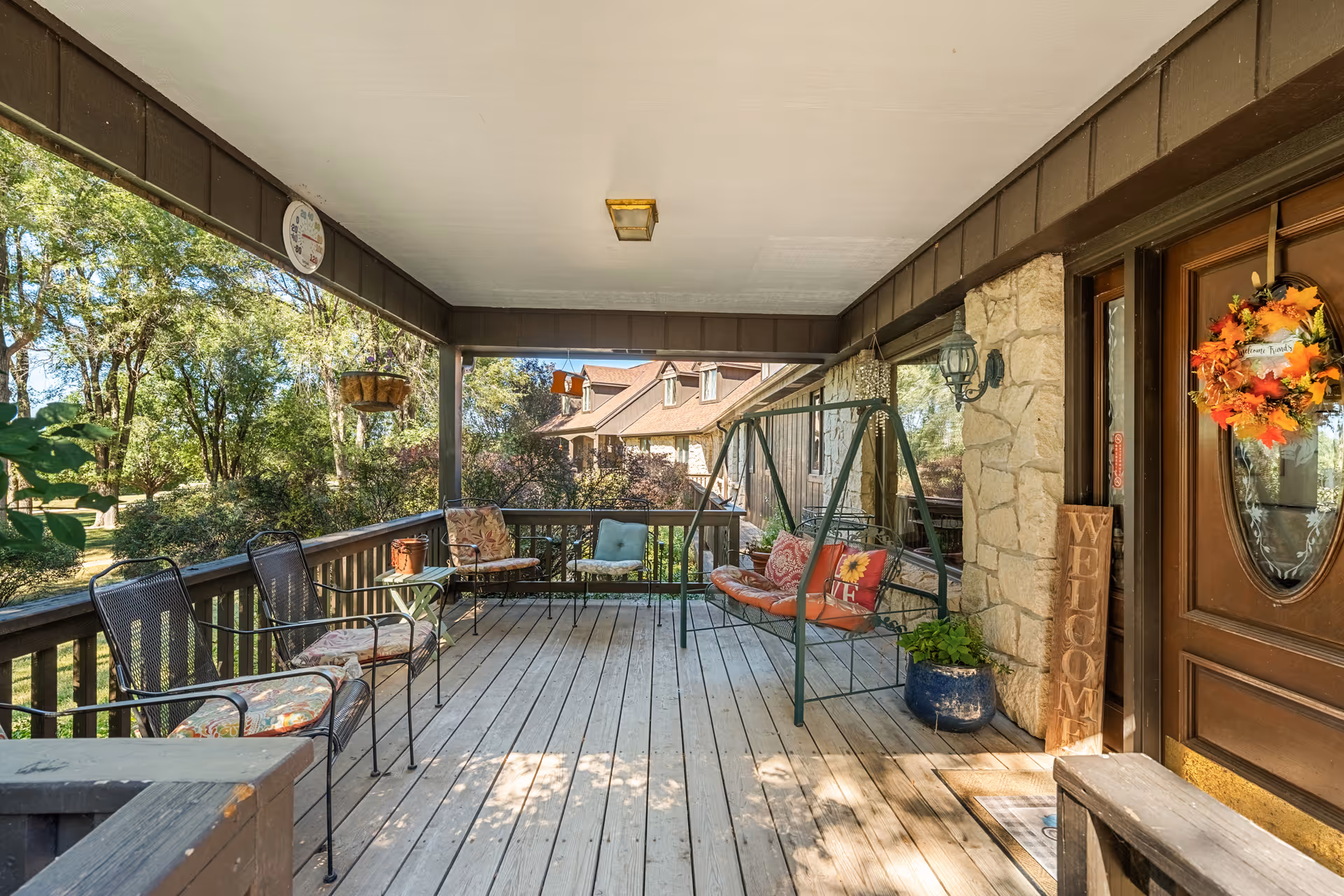 Covered wooden front porch with metal chairs, a cushioned swing, potted plants, and a wreath-decorated front door.