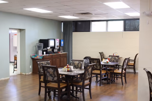 A dining area with several tables and chairs arranged neatly. Each table has condiments and napkins on it. Against one wall, there is a beverage station with a water dispenser and soda fountain. The room has wood flooring, light-colored walls, and windows near the ceiling letting in natural light.