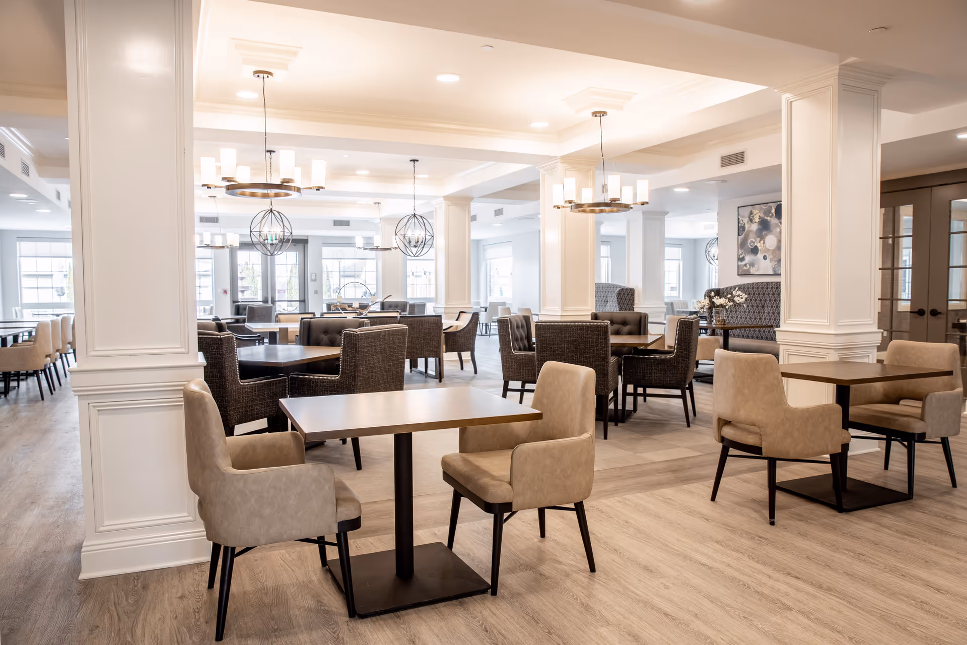 A bright and spacious dining room in a senior living facility with multiple tables and chairs arranged neatly. The room features modern light fixtures, large windows allowing natural light, white walls with decorative molding, and wood flooring.