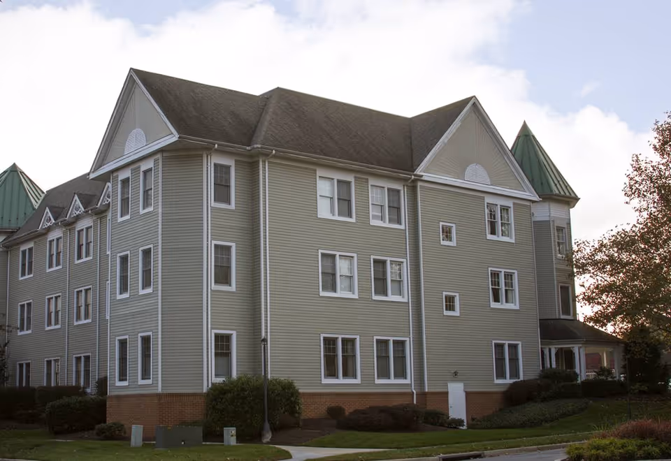 Exterior view of a three-story senior living facility building with light green siding, white trim, and a dark shingled roof. The building features multiple windows and a small covered entrance on the right side. There is a well-maintained lawn and bushes surrounding the building under a partly cloudy sky.