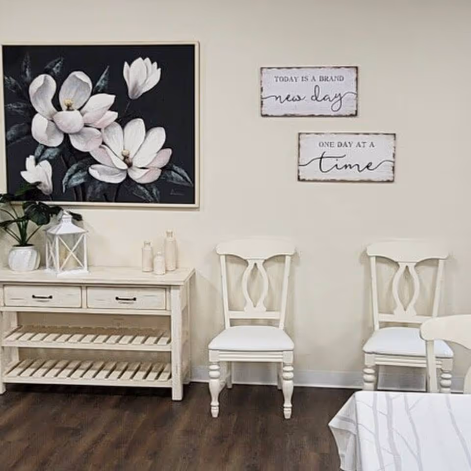 A cozy interior corner featuring two white wooden chairs with cushioned seats, a white wooden console table with two drawers and slatted shelves, decorative items including a potted plant, a white lantern, and small bottles. On the wall above are two framed inspirational signs reading 'TODAY IS A BRAND new day' and 'ONE DAY AT A time', alongside a large framed painting of white flowers with dark green leaves on a black background.