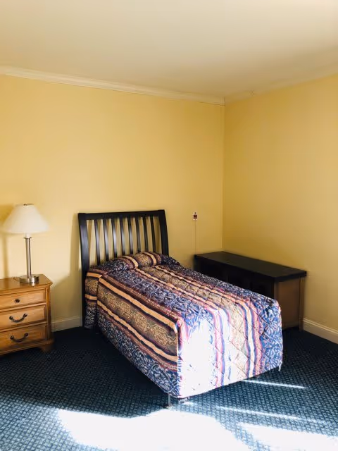 A simple bedroom with a single bed covered with a patterned quilt. The bed has a dark wooden headboard. To the left of the bed is a wooden nightstand with three drawers and a white table lamp. To the right of the bed is a dark wooden desk or table. The walls are painted light yellow and the floor is carpeted in a dark color. Sunlight is streaming into the room, casting shadows on the floor.