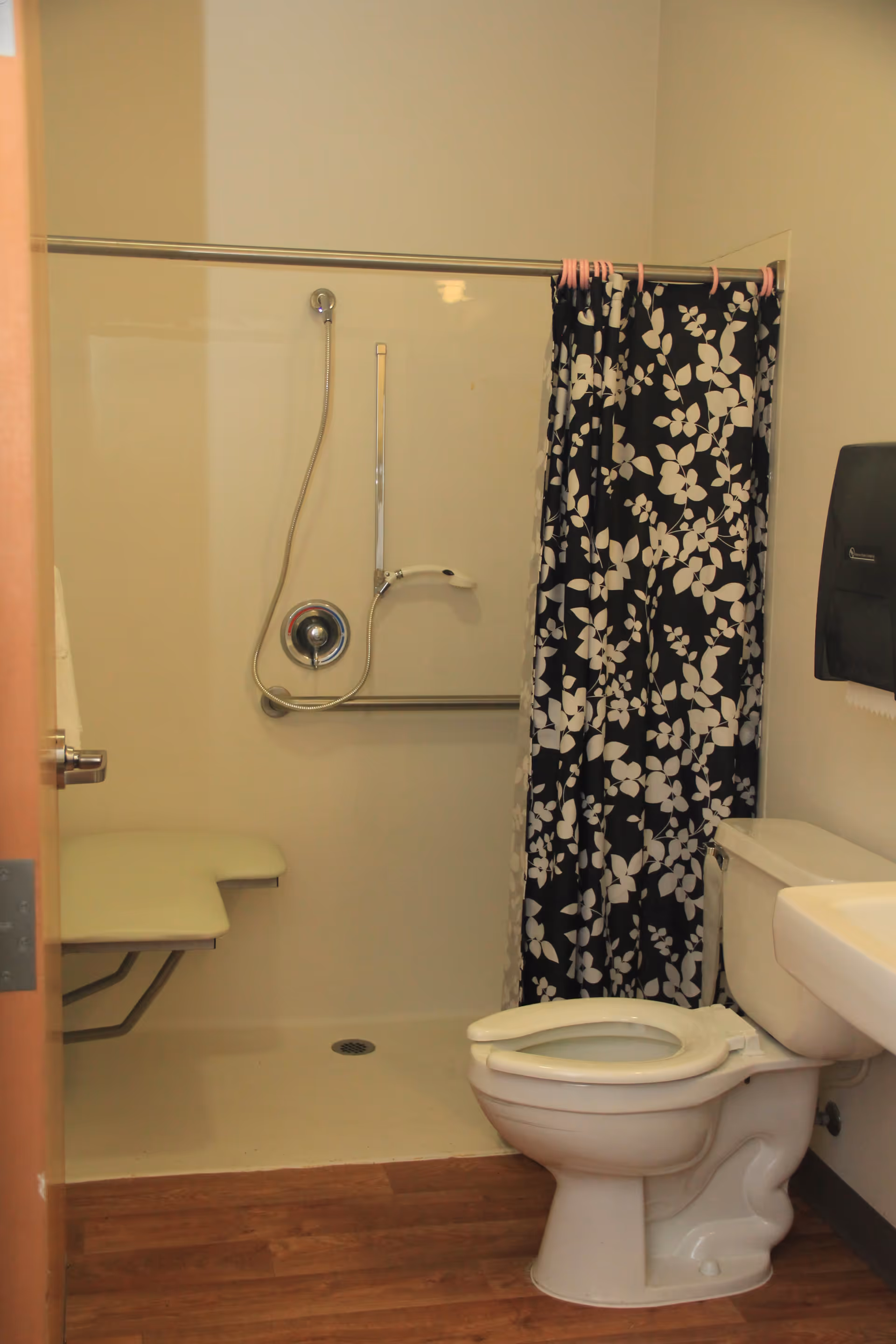 A bathroom with a toilet, a sink, and a walk-in shower area. The shower has a fold-down seat, a handheld showerhead, and a black and white floral shower curtain. The floor is wood-style, and there is a wall-mounted paper towel dispenser next to the sink.