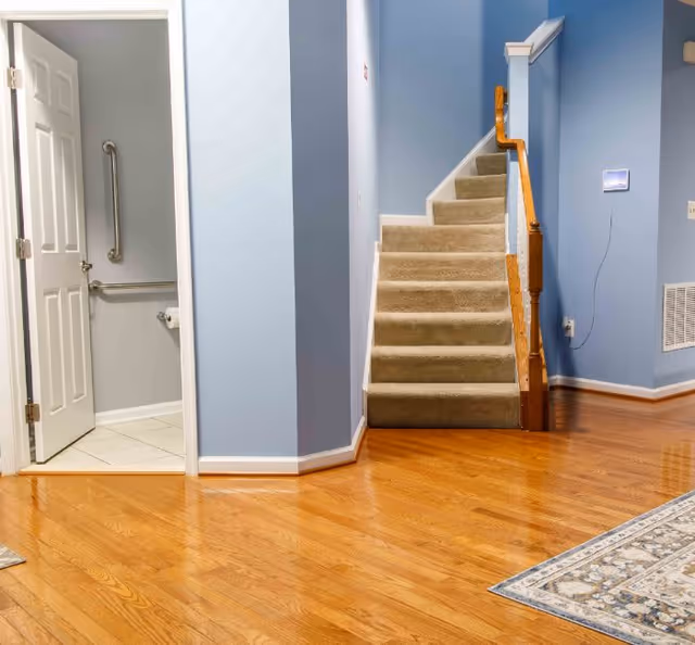Interior view of a hallway in an assisted living facility with light blue walls, wooden flooring, a carpeted staircase with a wooden handrail, and an open door leading to a bathroom equipped with grab bars and a toilet paper holder.