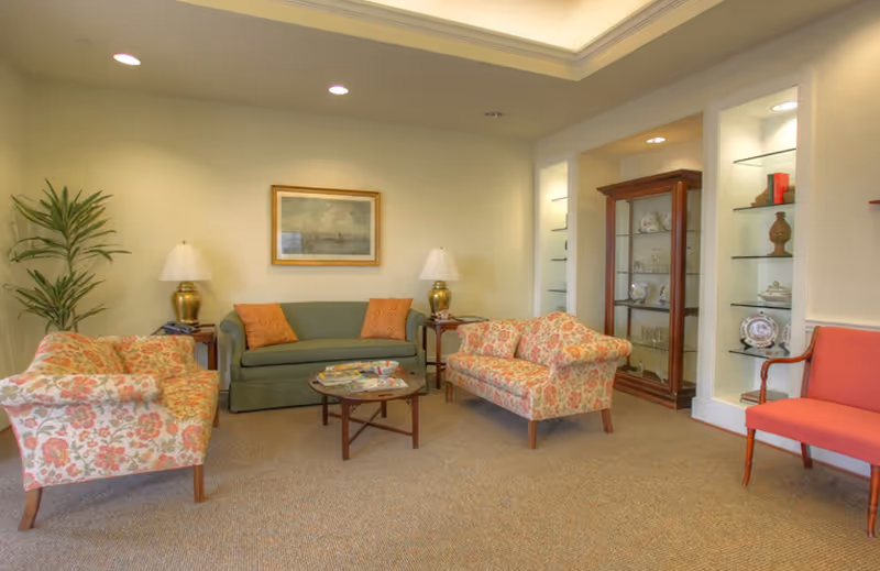 Communal sitting room with a green sofa, floral armchairs, coffee table, display cabinet and lamps.
