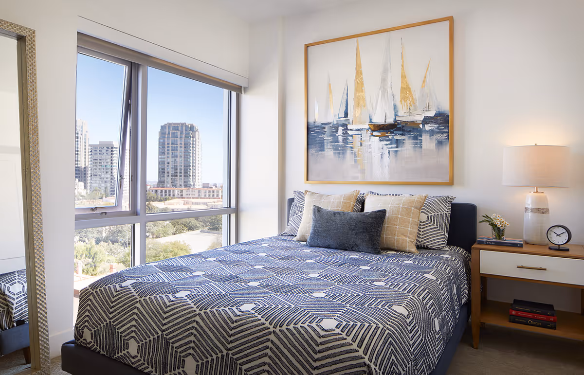 Bright modern bedroom with a large window showing city buildings, a bed with patterned bedding, sailboat artwork above the headboard, and a nightstand with a lamp.