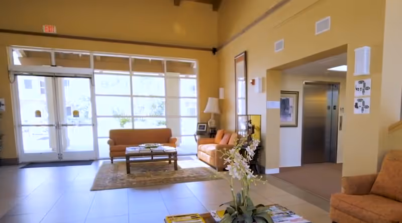 A bright senior living facility lobby with large glass doors and windows letting in natural light. The space features two orange sofas, a coffee table on a patterned rug, a side table with a lamp, and a plant in the foreground. An elevator and hallway are visible to the right.