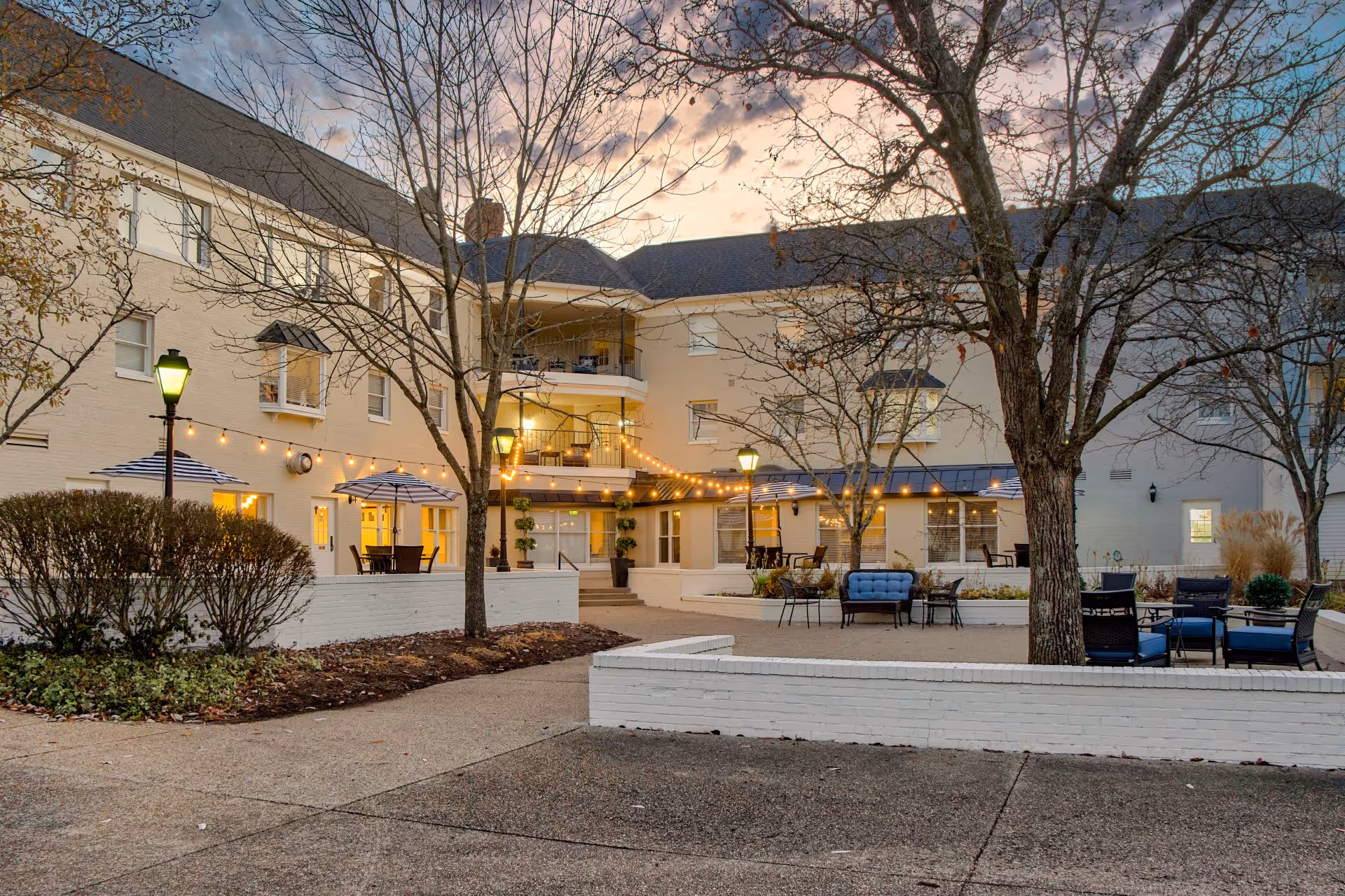 Courtyard and outdoor seating area of a multi-story senior living building illuminated with string lights at dusk.
