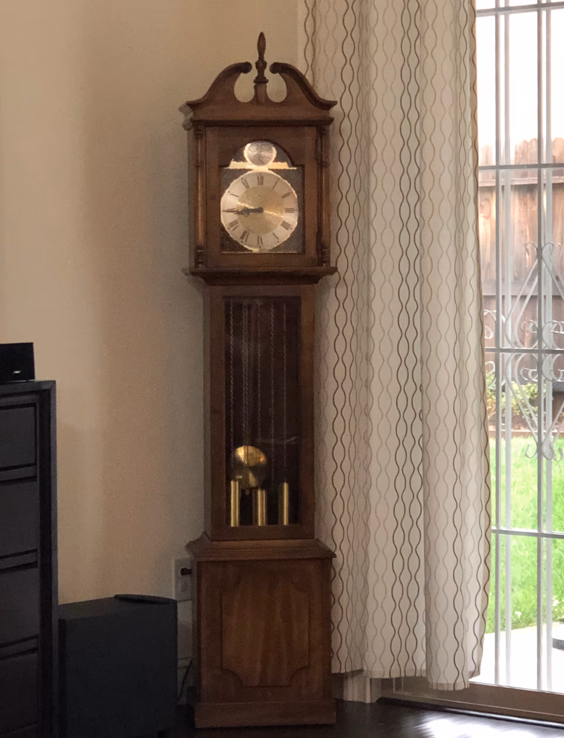 A wooden grandfather clock standing next to patterned curtains and a sliding glass door in a living room.