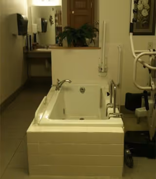 A bathroom area featuring a white tiled bathtub with a faucet and grab bars, a plant on a ledge behind the tub, a sink with a mirror and soap dispenser in the background, and a piece of exercise equipment to the right.