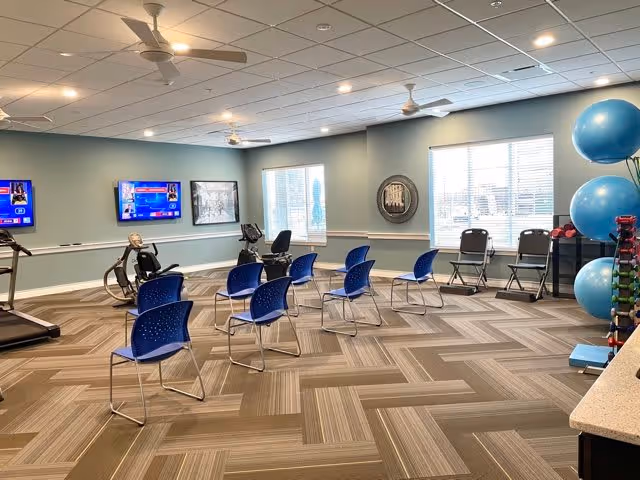A spacious exercise room with several blue chairs arranged in rows facing two wall-mounted televisions. The room has large windows letting in natural light, exercise equipment including stationary bikes, and a rack of large blue exercise balls and dumbbells. The walls are painted light blue with white trim, and the ceiling has recessed lighting and ceiling fans.