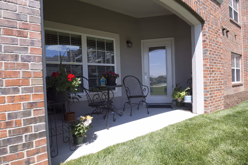 A covered ground-floor patio with wrought-iron chairs, a small table and potted plants outside a brick building.