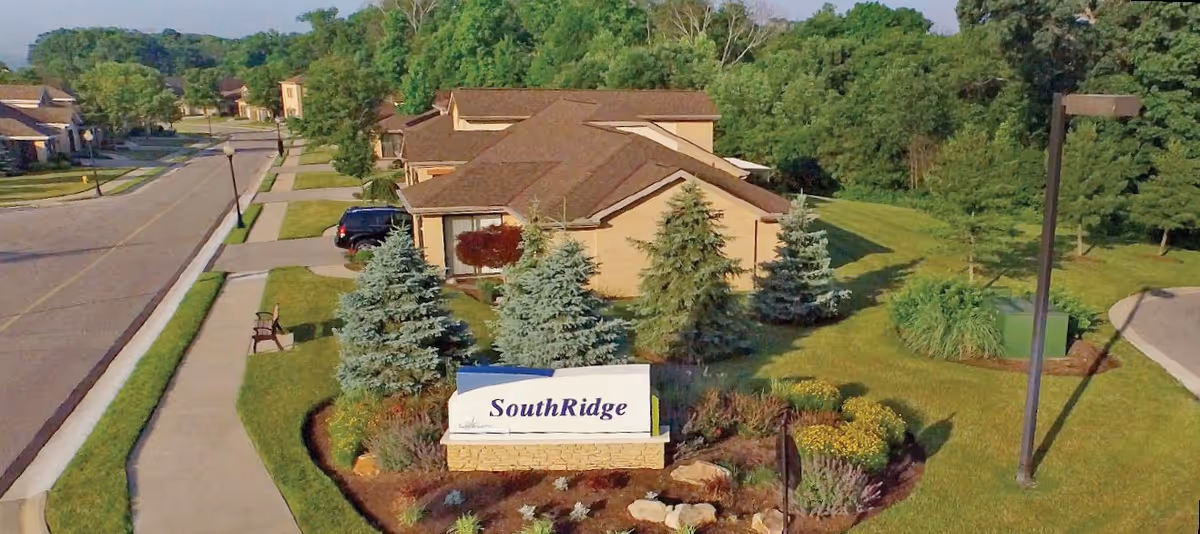 Aerial view of a residential community with single-story houses, well-maintained lawns, trees, and a sidewalk. A sign in the foreground reads 'SouthRidge'.