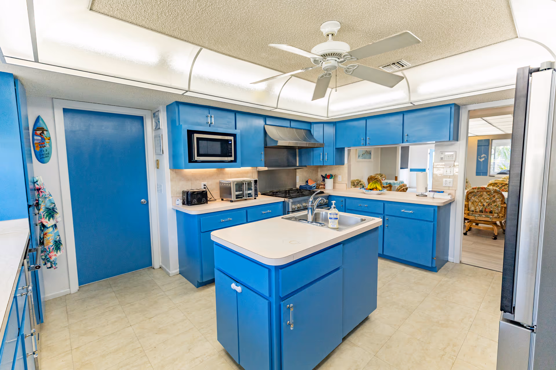 Bright blue kitchen featuring a central island with sink, ceiling fan, microwave, stovetop with hood, and stainless refrigerator.