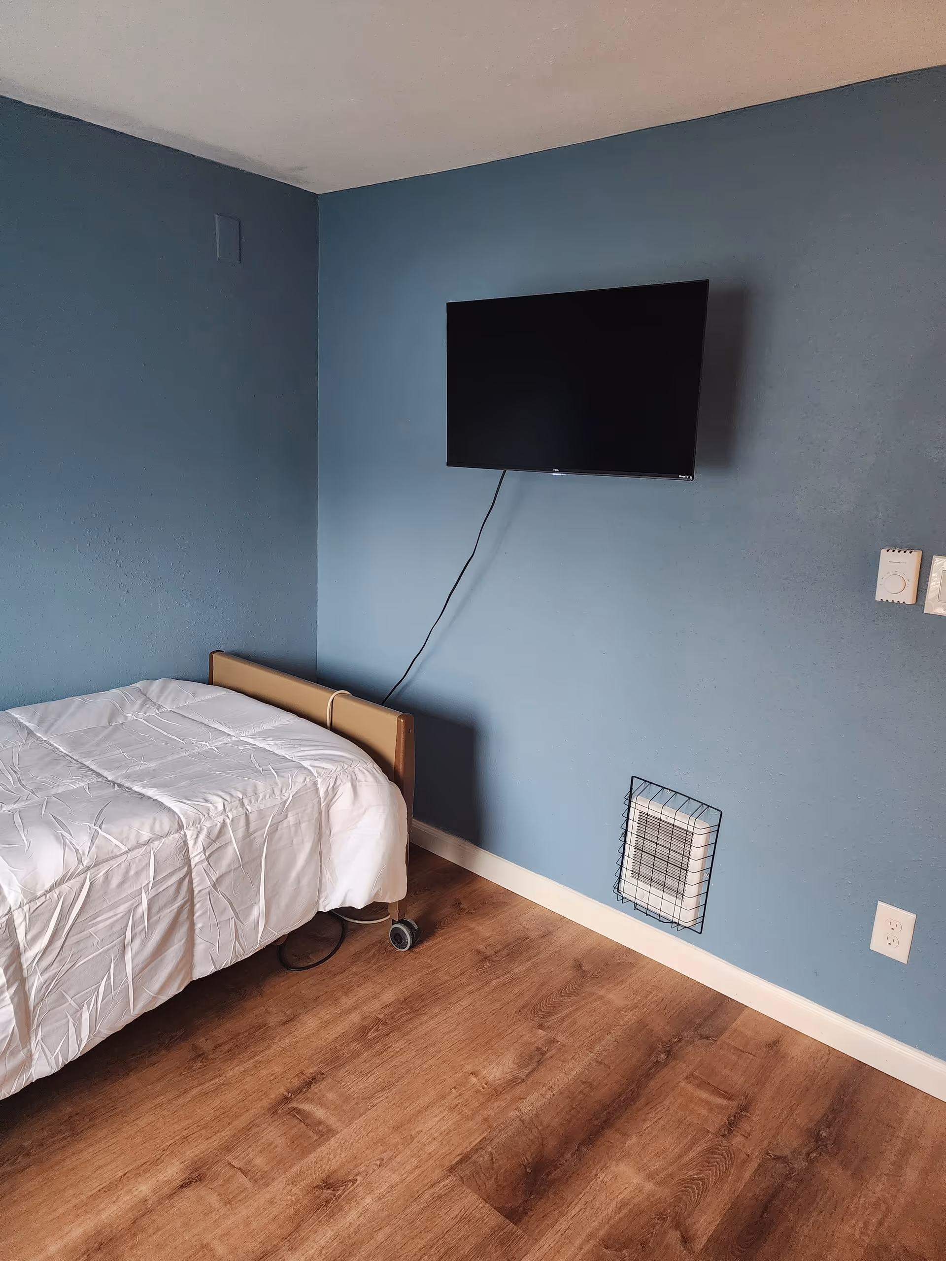 A simple bedroom with a single bed covered with a white comforter, a wall-mounted flat-screen TV, and a blue painted wall with a vent and electrical outlets. The floor is wooden.