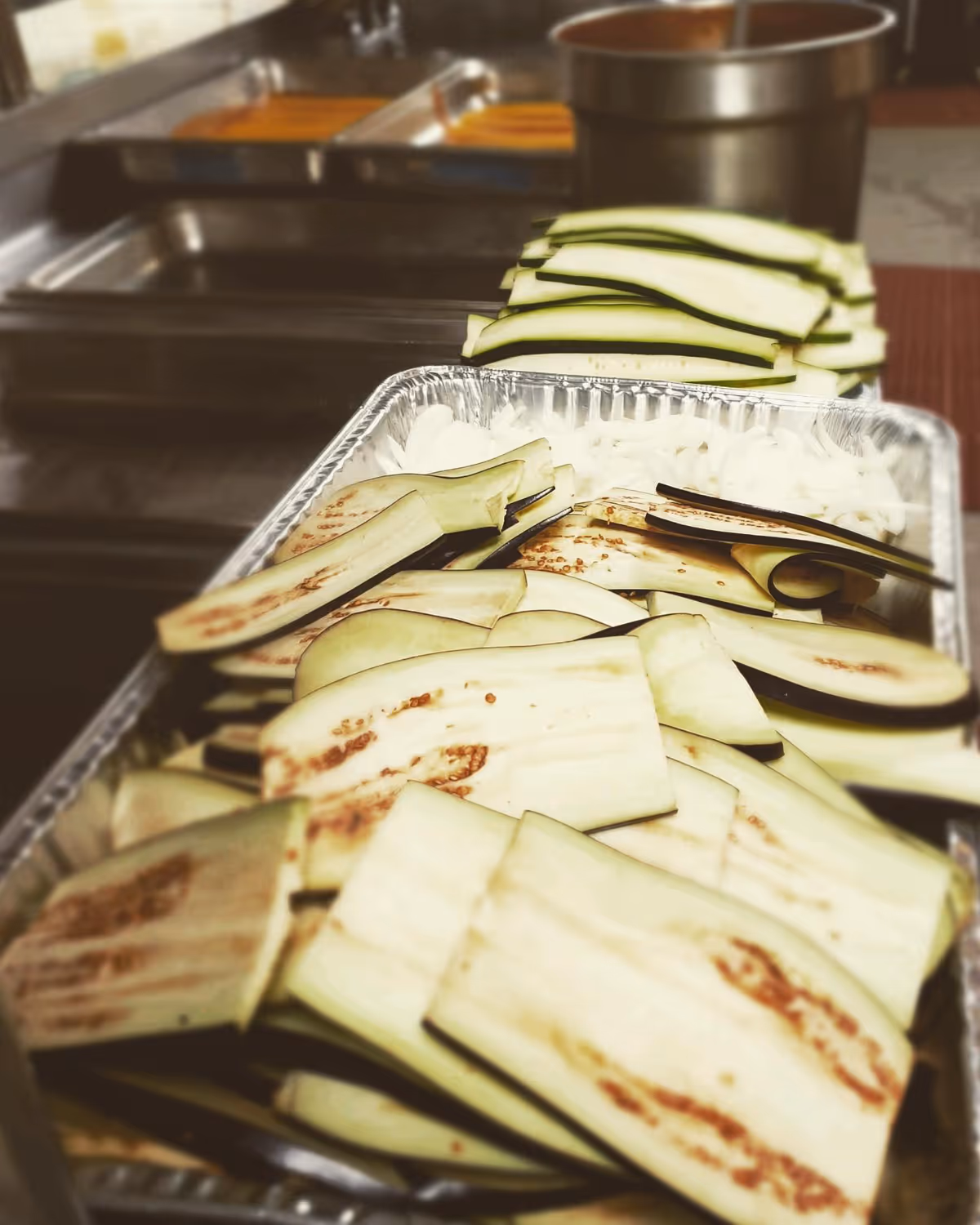 Close-up view of sliced eggplants and zucchini arranged in a metal tray on a kitchen counter with a pot and other kitchen utensils in the background.