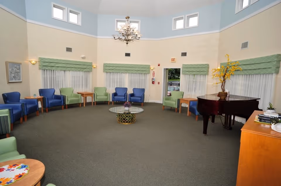 A spacious, circular common area with a mix of blue and green armchairs arranged around the perimeter. The room features large windows with green valances and sheer white curtains, a glass coffee table in the center, a grand piano with a vase of yellow flowers on top, and a wooden cabinet with books and plants. The walls are painted beige with a light blue upper section, and there is a chandelier hanging from the ceiling.