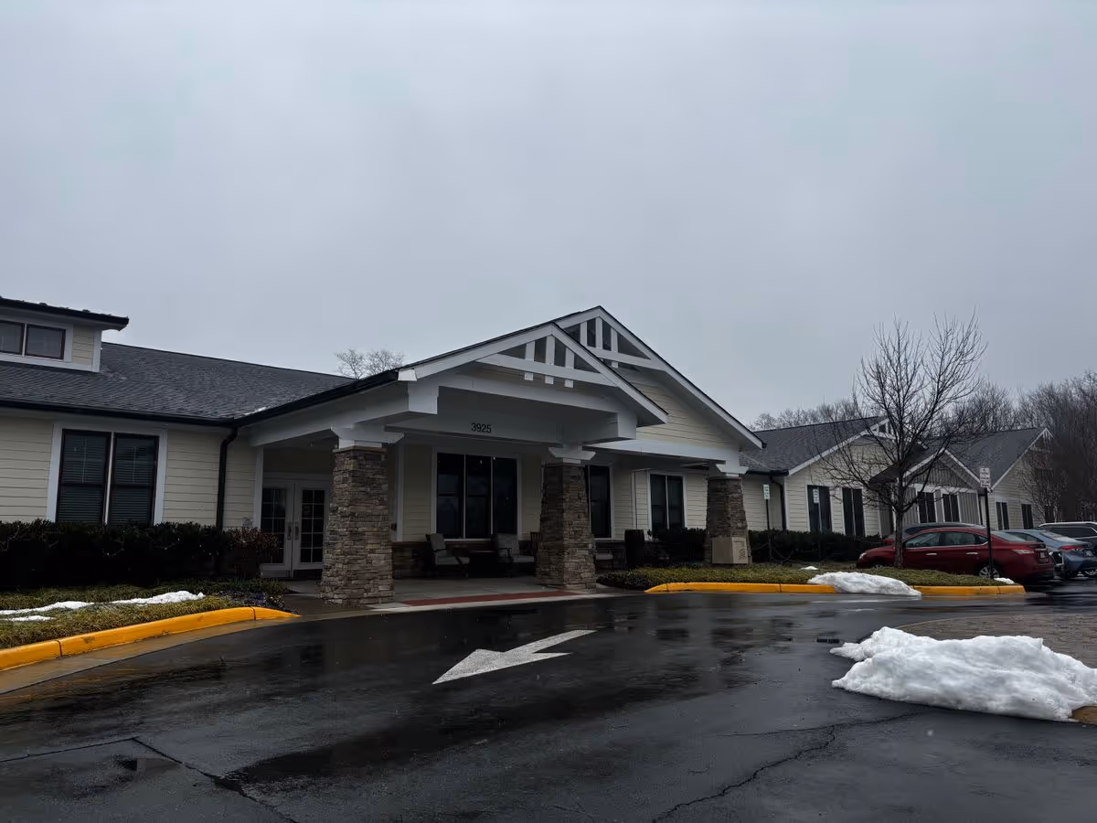 Covered entrance of a single-story memory care building with stone columns, a wet driveway with directional arrows, parked cars, and patches of melting snow.