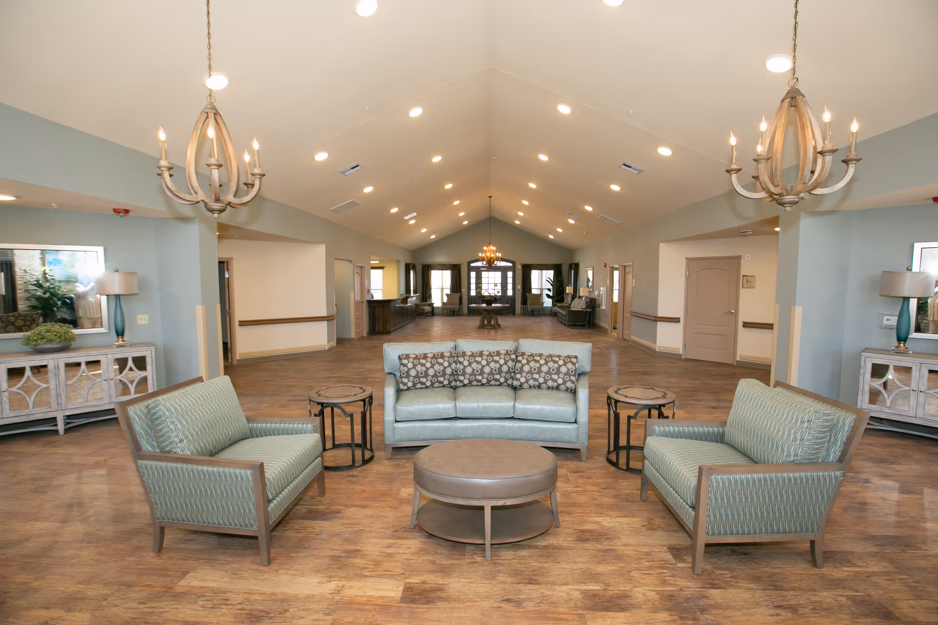 A spacious and well-lit common area in a senior living facility featuring a seating arrangement with a light blue sofa, two matching armchairs, and a round coffee table in the center. The room has wooden flooring, light blue walls, two chandeliers hanging from the ceiling, and side tables with lamps and decorative plants on either side.