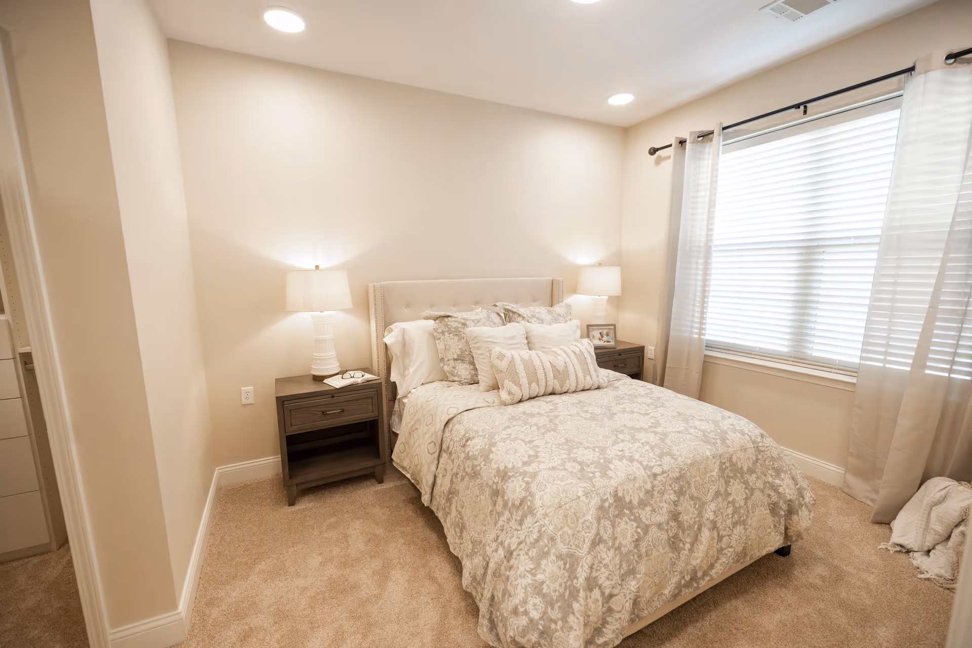 A cozy bedroom with a neatly made bed featuring patterned bedding and multiple pillows. There are two bedside tables, each with a lamp, one holding a pair of glasses and a book, and the other a framed photo. A large window with white blinds and light curtains lets in natural light. The room has beige walls and carpeted flooring.