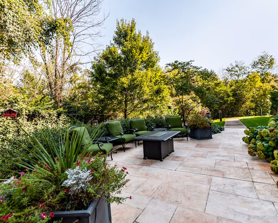 Outdoor patio area with green cushioned chairs arranged around a rectangular fire pit table, surrounded by lush greenery and trees under a clear sky.