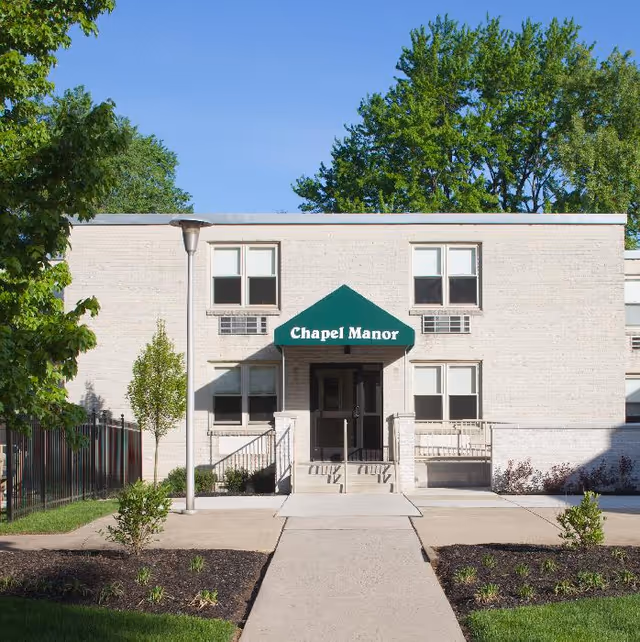 Front exterior view of a two-story light-colored brick building with a green awning that reads 'Chapel Manor'. There are windows on both floors and a concrete walkway leading to the entrance. The area is landscaped with small trees, plants, and a lamp post, with a clear blue sky and large trees in the background.