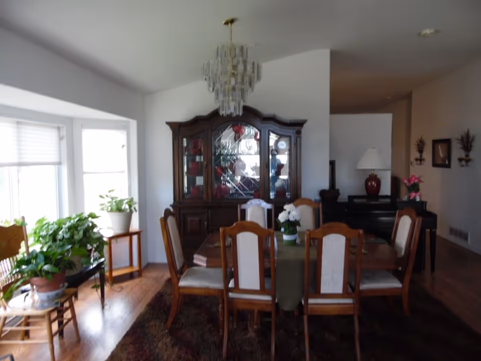 A dining room with a wooden table and six chairs under a chandelier, a china cabinet against the wall, potted plants by a bay window and a lamp on a side table.