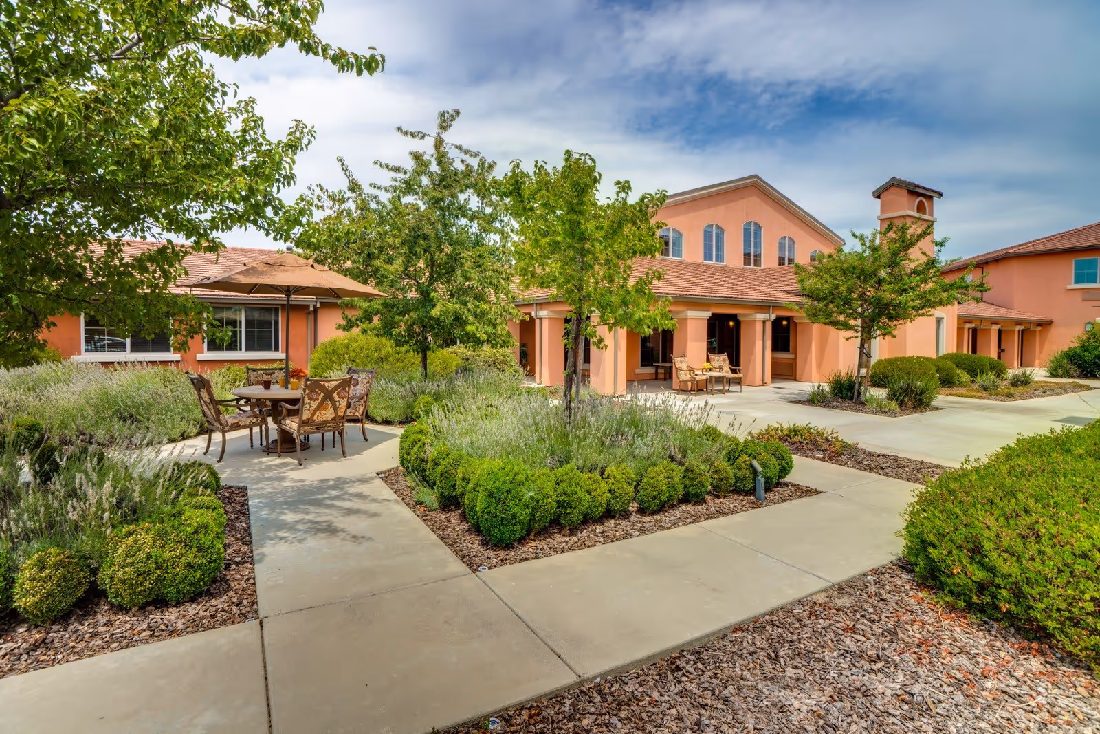 Outdoor courtyard area at Eskaton Village Roseville featuring a paved walkway, green shrubs, trees, and a seating area with a table and chairs under a large umbrella. The building in the background has a peach-colored exterior with multiple windows and a covered patio with additional seating.