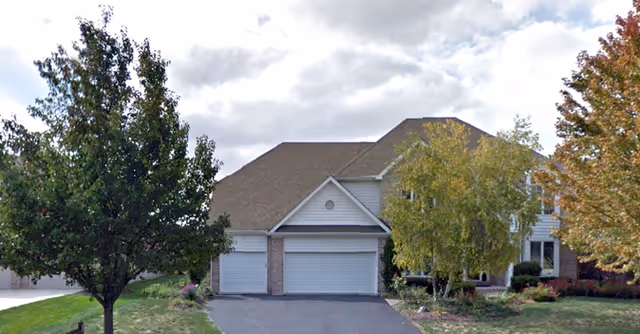 A two-story residential building with a three-car garage, surrounded by trees and landscaping under a cloudy sky.