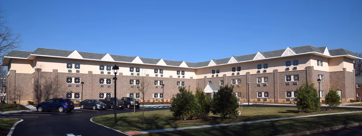 Exterior view of a large, three-story senior living facility building with brick and beige walls, multiple windows, a parking lot with several cars, and a landscaped lawn with small trees and a clear blue sky.